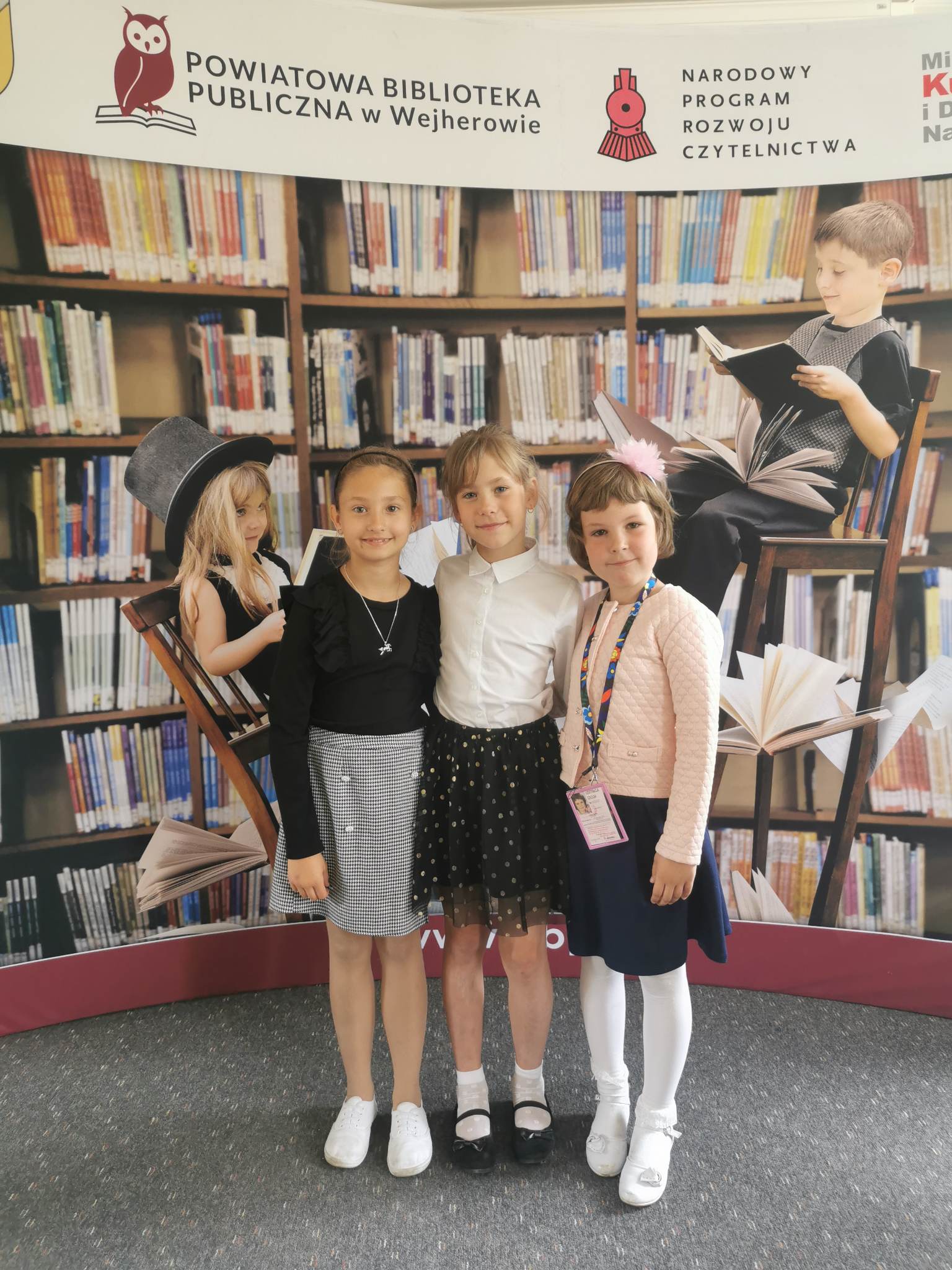 Zdjęcie trzech uczennic, które reprezentowały naszą szkołę w konkursie na tle pięknej gazetki przedstawiającej dziewczynkę i chłopca, którzy siedzą na krzesłach i czytają oraz książek.
