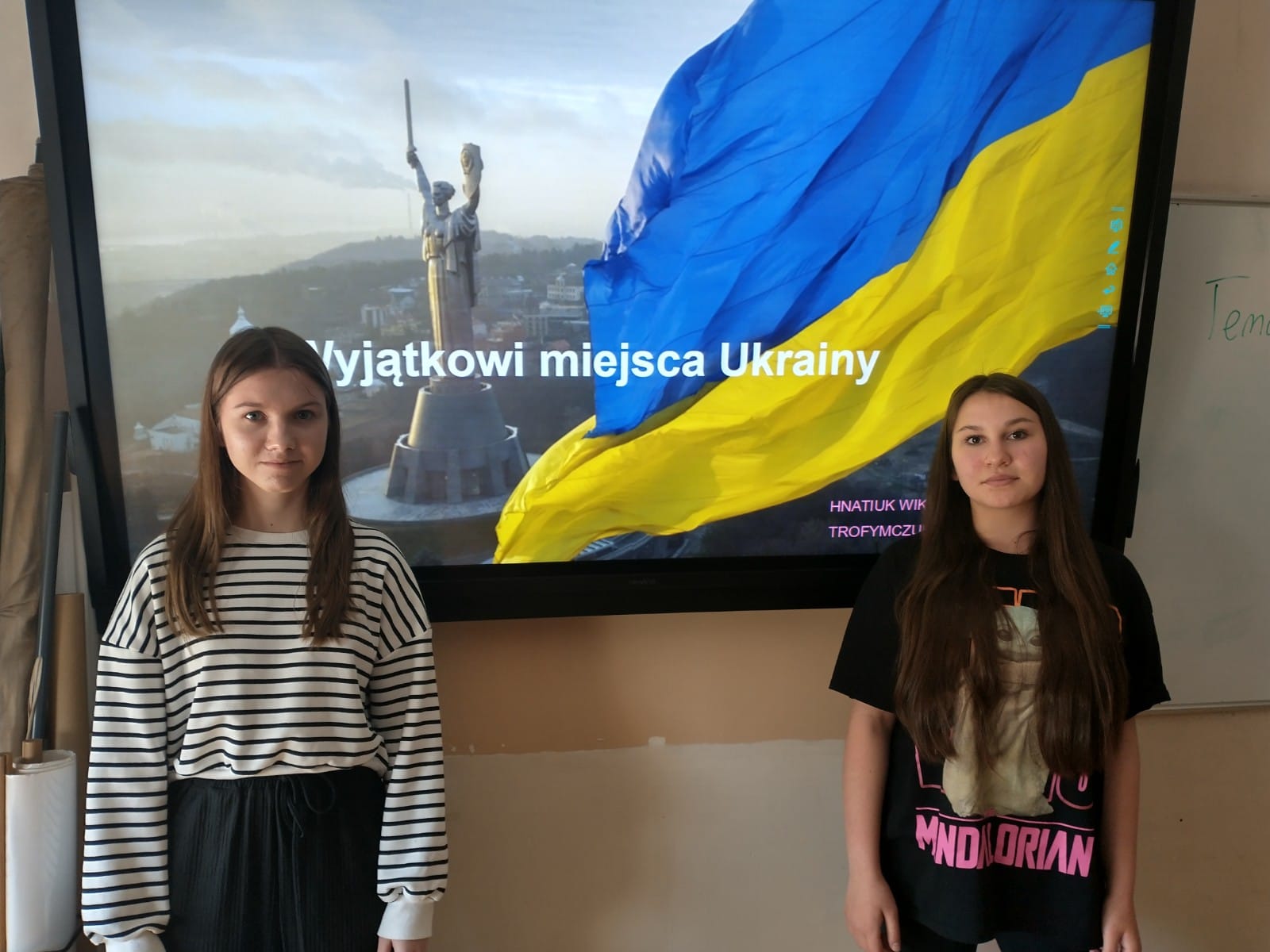 Dwie uczennice klasy 7b stoją na tle prezentacji multimedialnej o wyjątkowych miejscach Ukrainy.