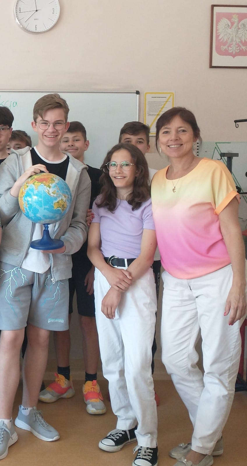 Nauczycielka geografii stoi wraz z uczniami w sali lekcyjnej.