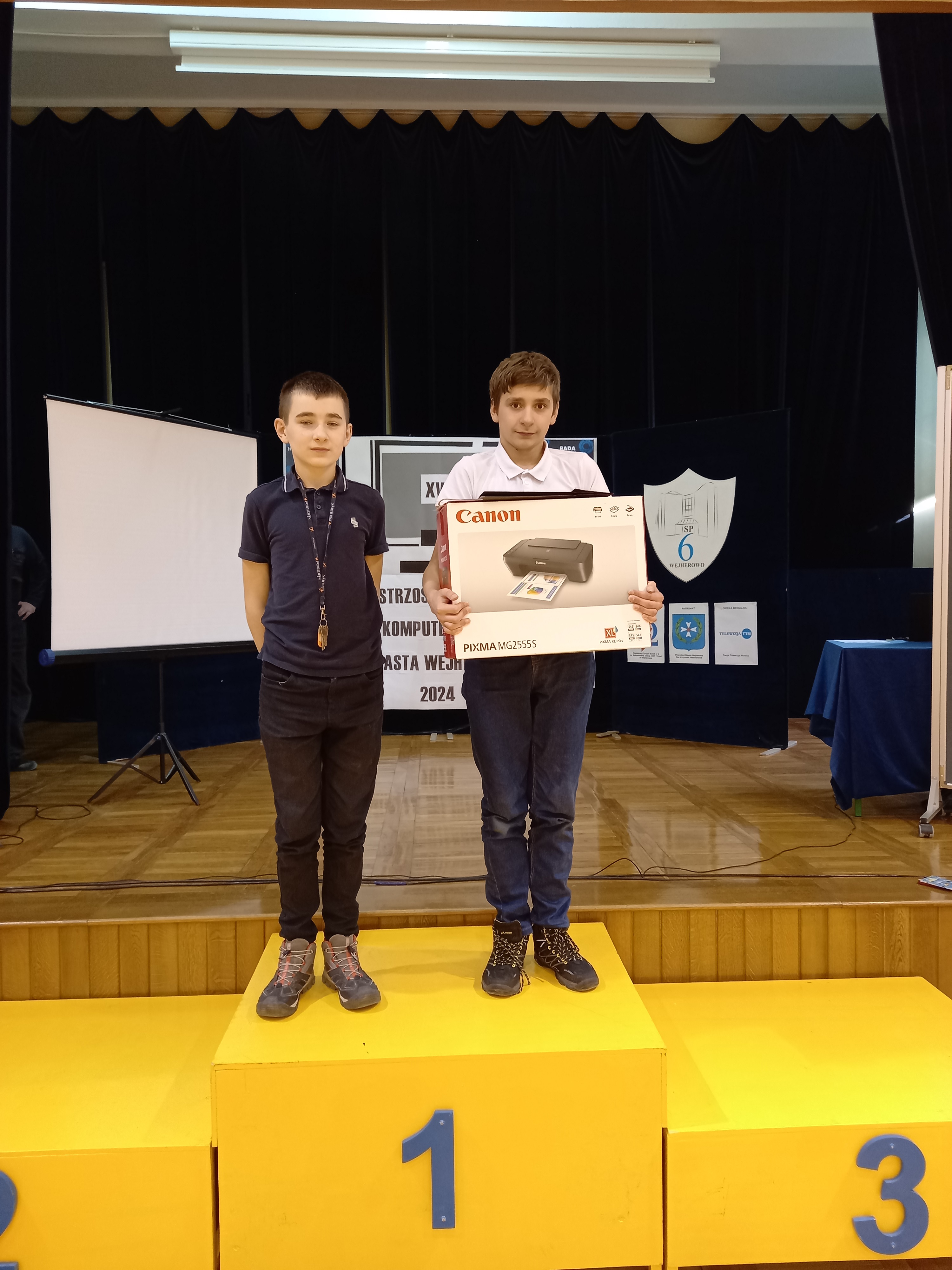 Dwóch chłopców na podium - 1 miejsce. Na szyi jednego medal, drugi trzyma w rękach karton z nagrodą - drukarką.