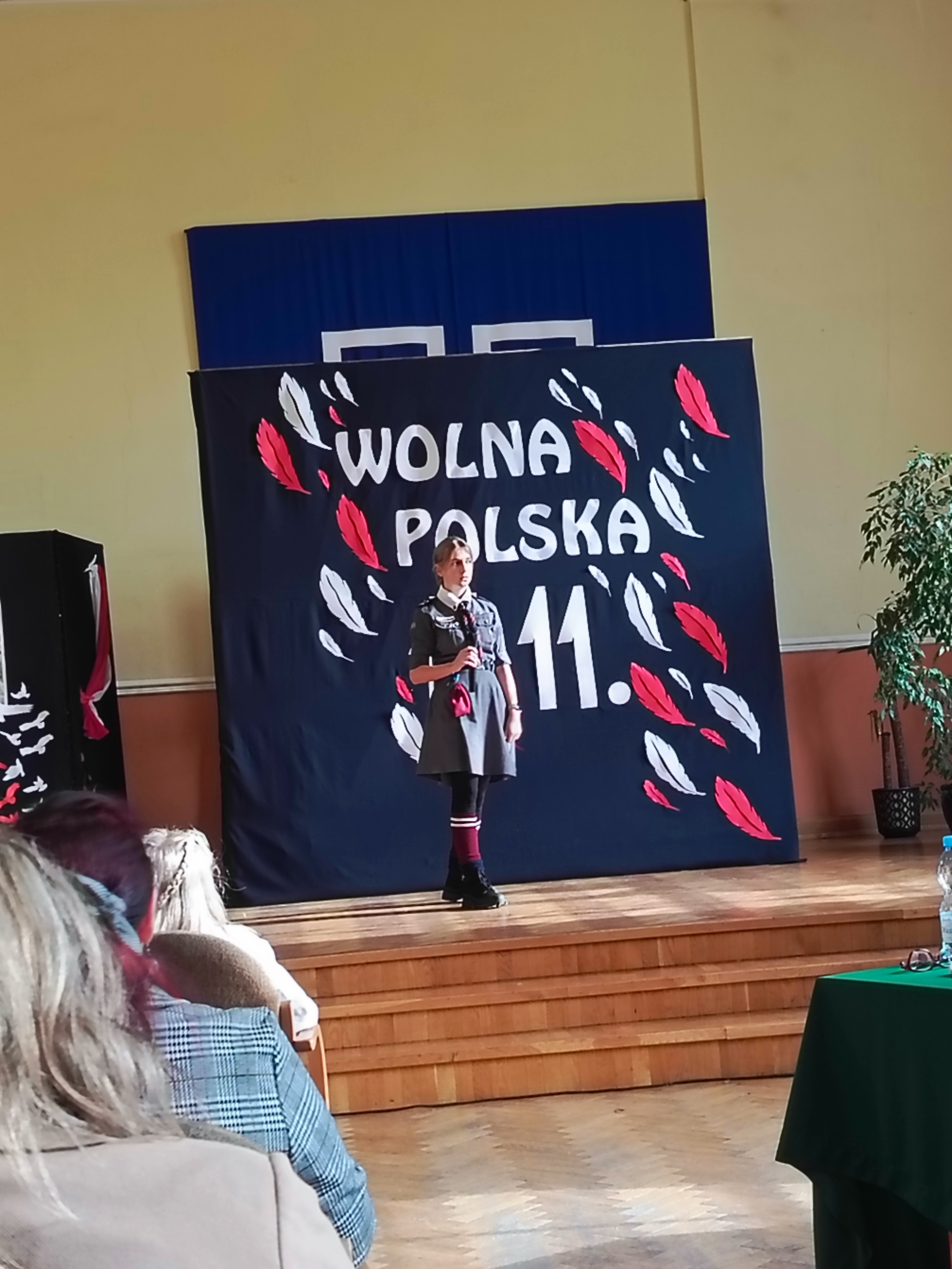 Na scenie dziewczynka z mikrofonem ubrana w harcerski mundur. W tle tablica z napisem: 11.11 Wolna Polska
