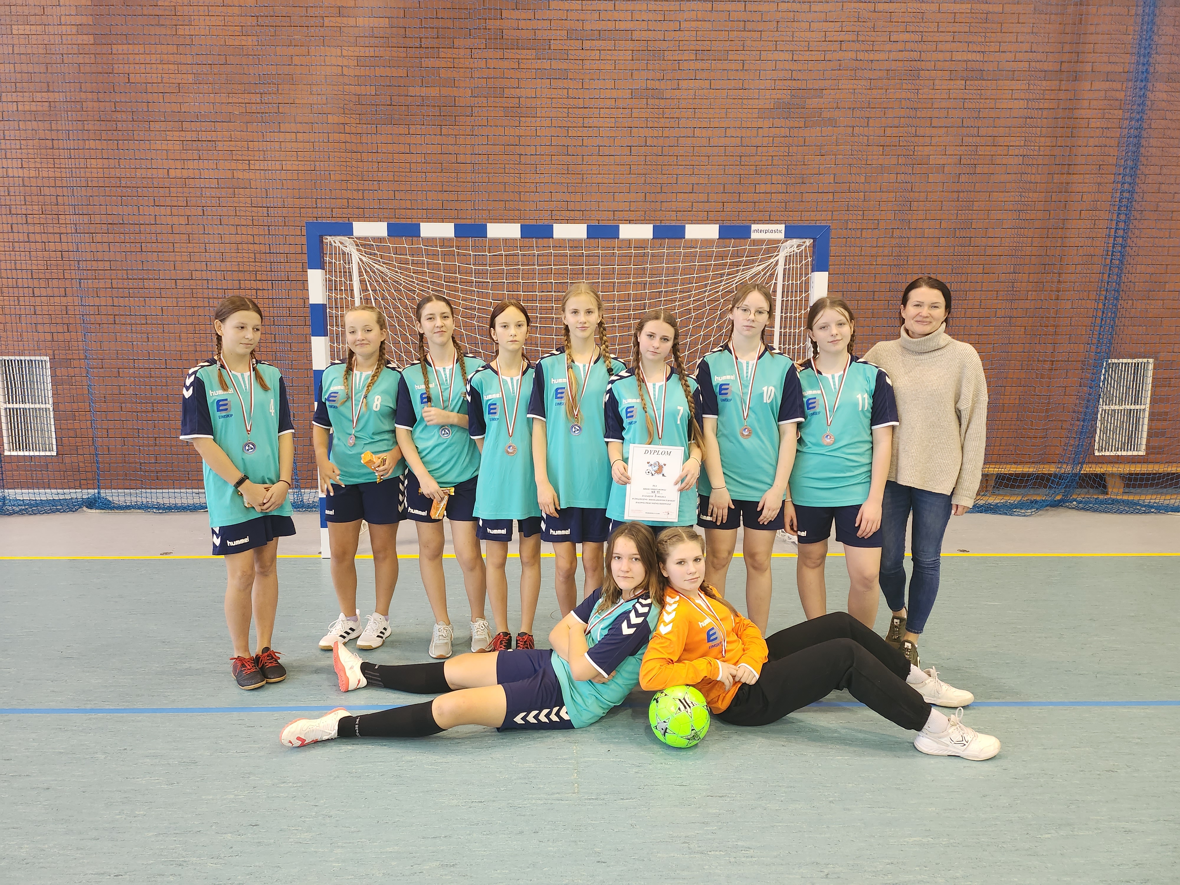 Zdjęcie przedstawia reprezentację dziewcząt SP 11 w piłce nożnej halowej wraz z trenerem w turkusowych strojach i z medalami.