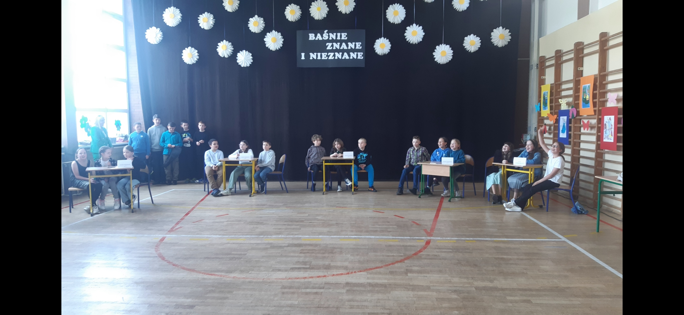 Na sali gimnastycznej, przy stolikach siedzą drużyny biorące udział w zawodach. Z tyłu napis Baśnie znane i nieznane.