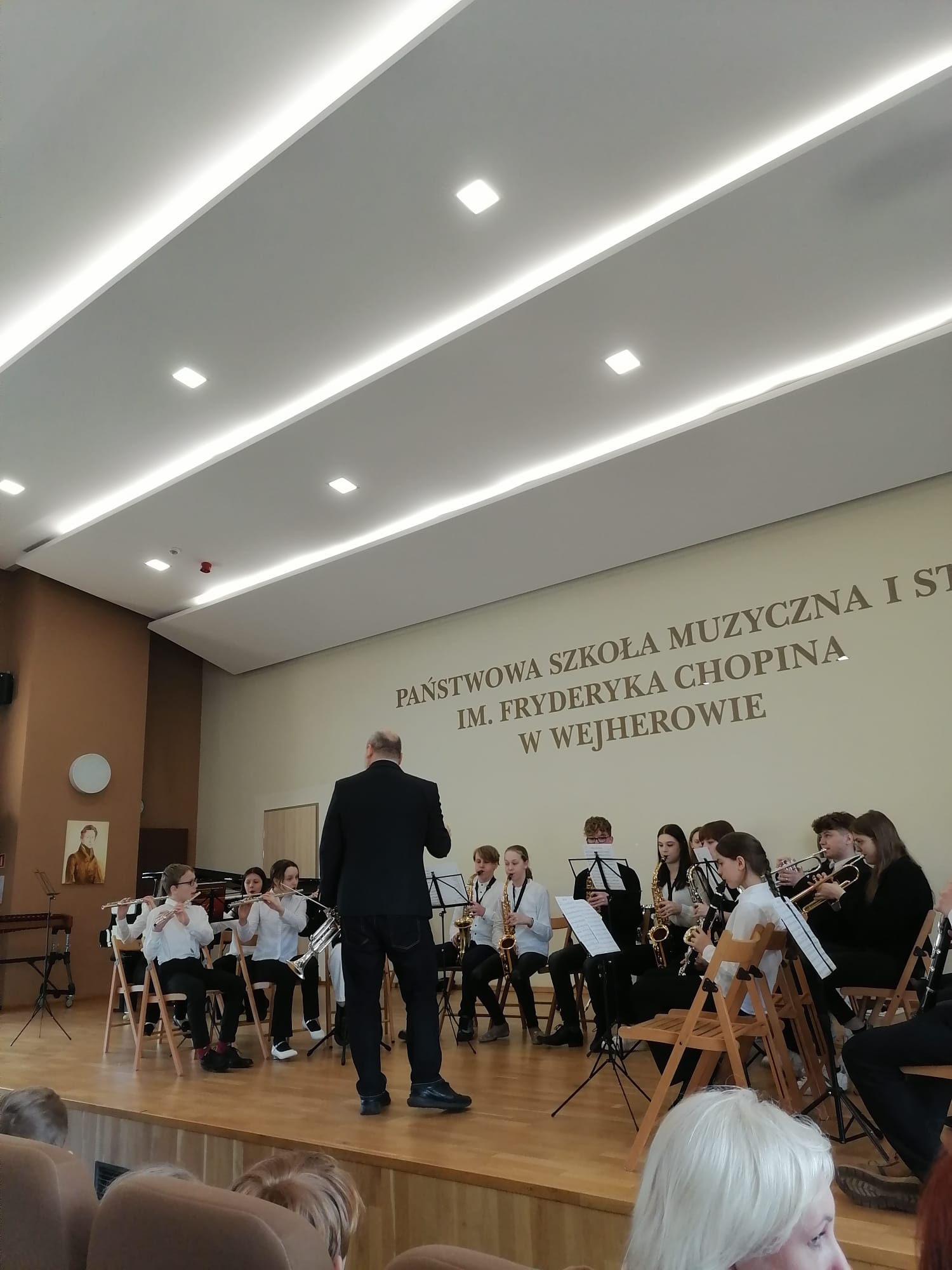 Na podeście w auli Szkoły Muzycznej siedzą uczniowie grający na różnych instrumentach w orkiestrze, na środku stoi dyrygent