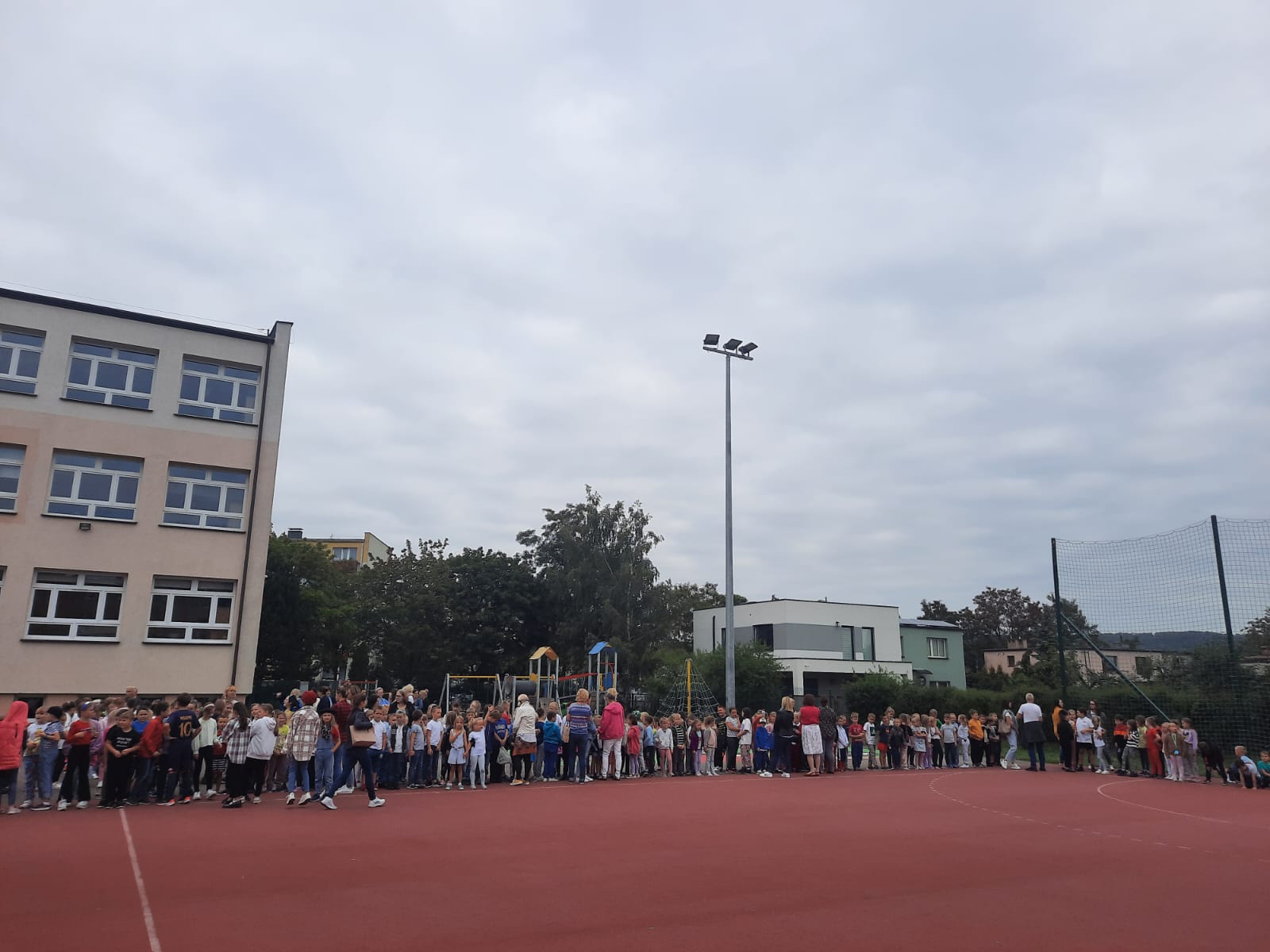 Osoby obecne w szkole podczas ogłoszenia próbnego alarmu przeciw pożarowego stoją na boisku szkolnym.