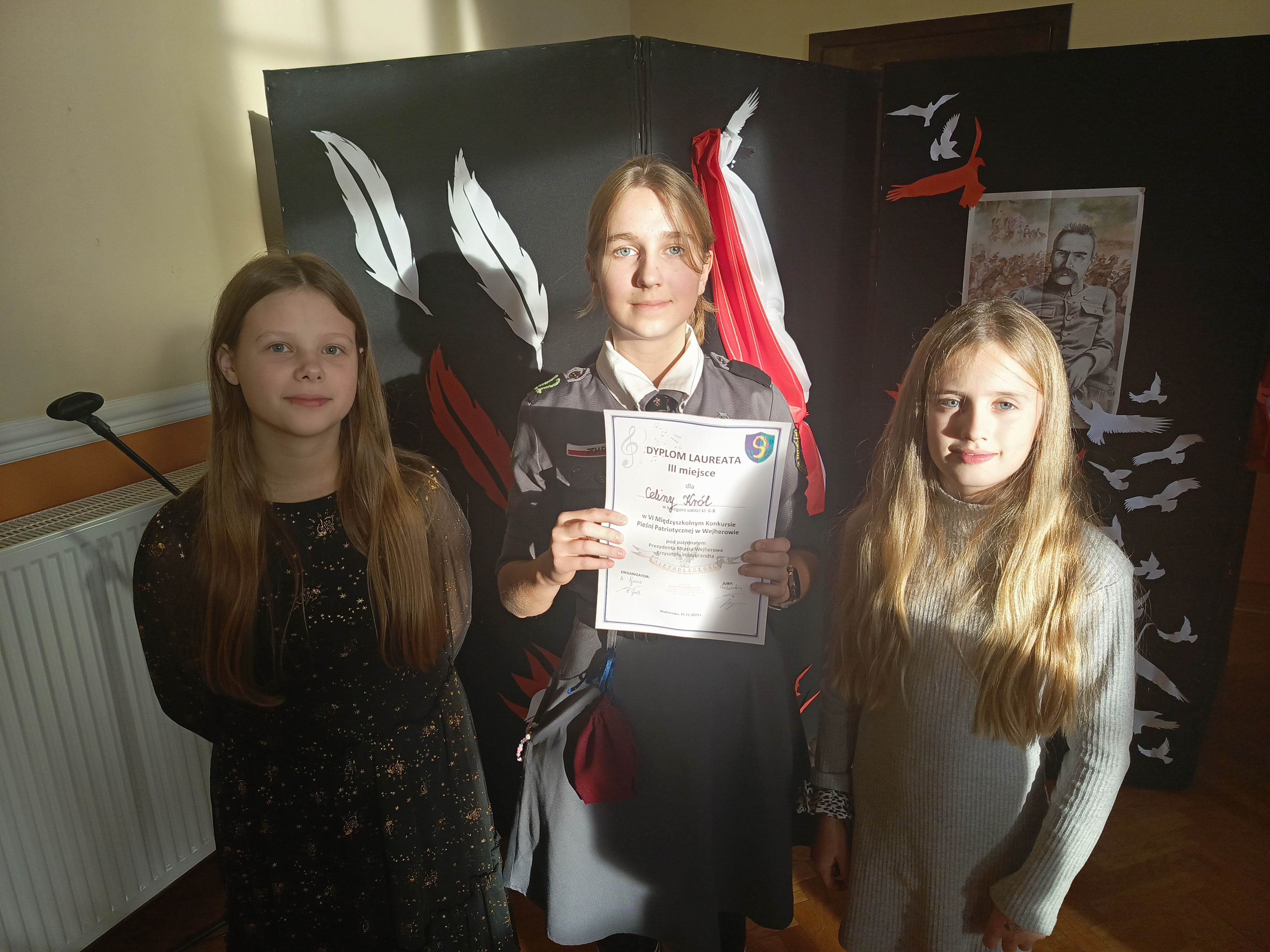 Na zdjęciu trzy dziewczynki, jedna z nich trzyma dyplom. W tle granatowa tablica na niej polska flaga i wydruk  zdjęcia Marszałka Piłsudskiego