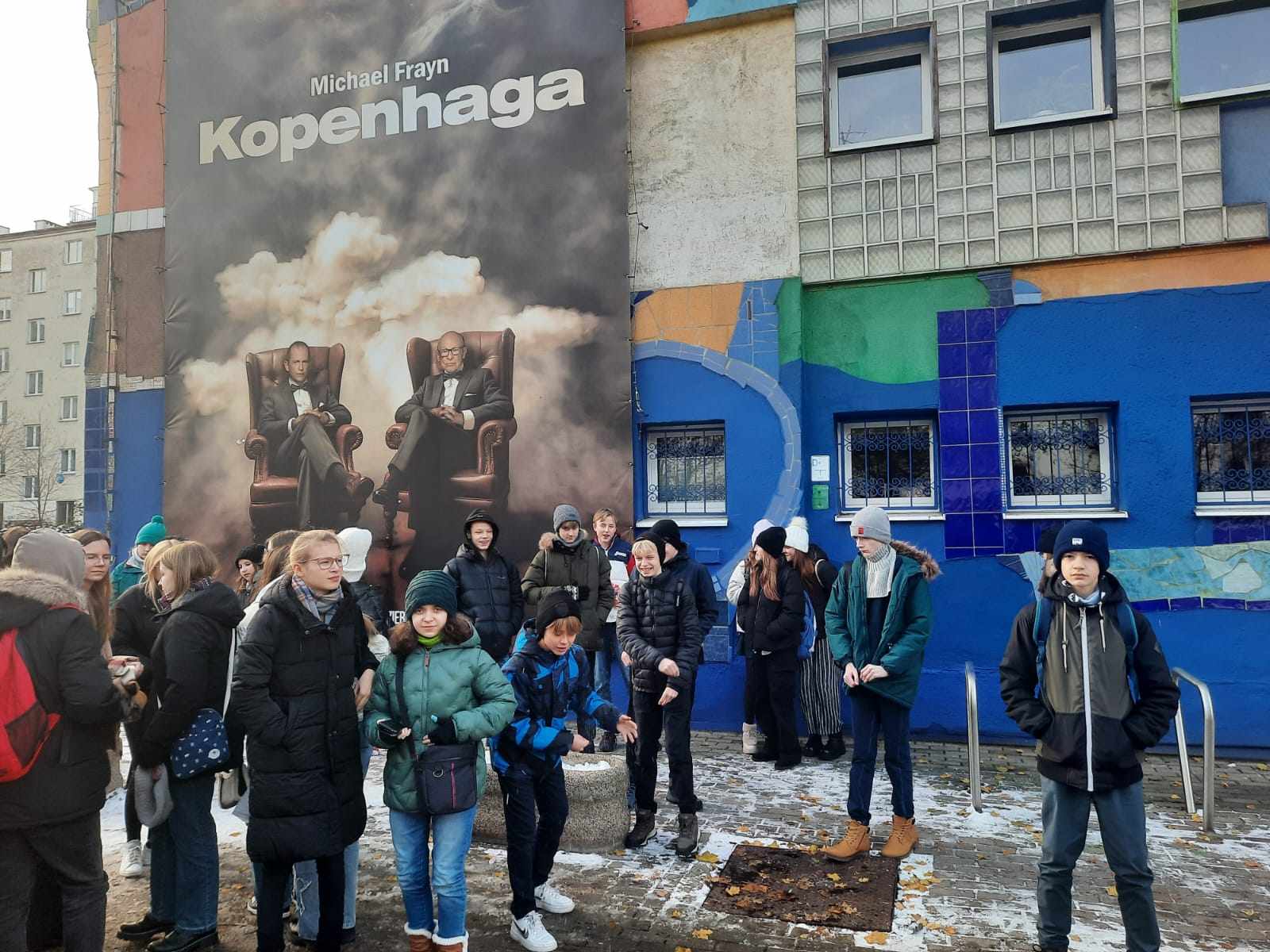 Grupa młodzieży przed budynkiem. Ściana budynku pokryta graffiti. Na ścianie wisi plakat promujący spektakl, napis: Michael Frayn "Kopenhaga".