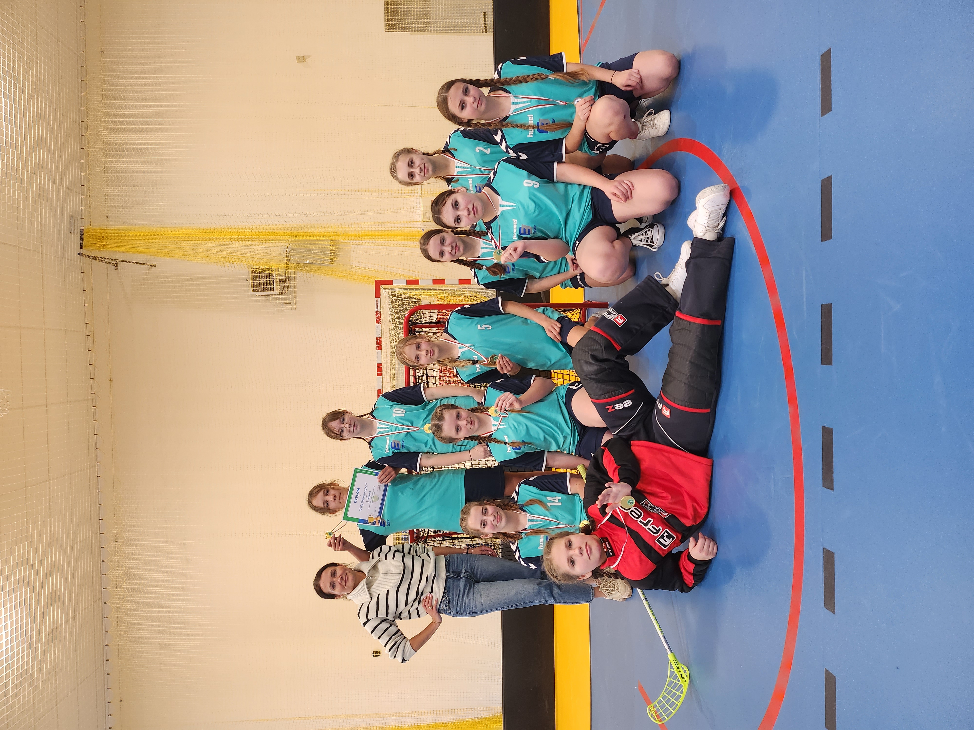 Drużyna dziewcząt na sali gimnastycznej. 10 uczennic w turkusowych strojach stoi razem z trenerką. Na szyjach i w rękach złote medale.