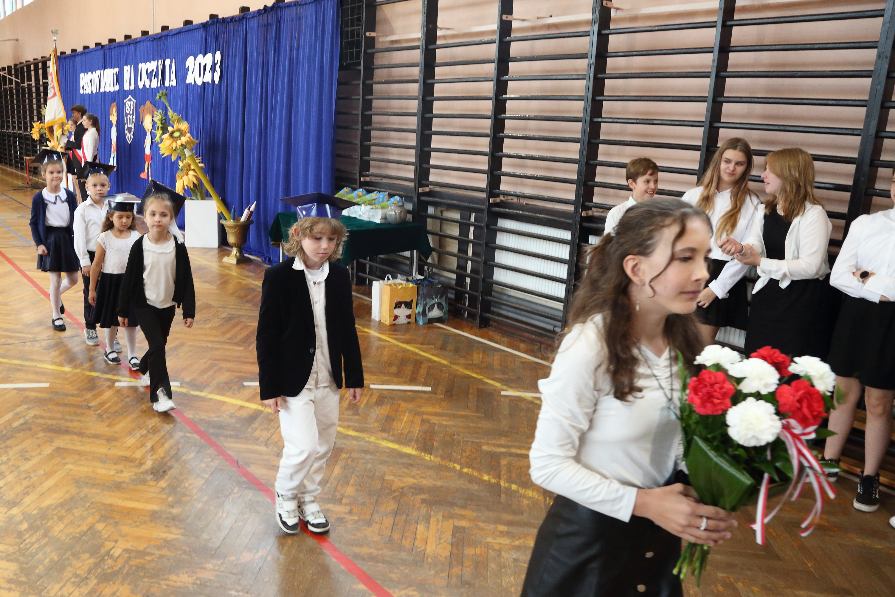 Na zdjęciu widać uczennicę klasy ósmej trzymającą bukiet złożony z białych i czerwonych goździków, za nią w rzędzie delegaci klas pierwszych do łożenia kwiatów pod tablicą patrona szkoły.